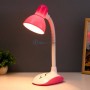 Lampe de bureau rose à col flexible allumée posée sur une table avec un petit pot de plante et un cadre en arrière-plan.