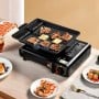 Réchaud à gaz noir avec plaque de cuisson grill, viande et légumes en cuisson, posé sur une table avec assiettes et couverts.