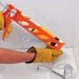 Pistolet à silicone orange tenu par une personne gantée appliquant du silicone dans un coin carrelé au-dessus d'un lavabo.