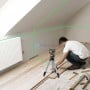 Niveau laser vert posé sur un trépied, projetant deux lignes sur un mur et un plafond, homme posant un parquet au sol